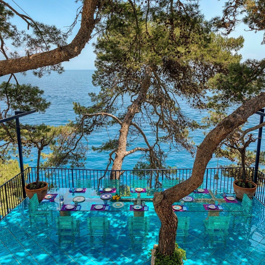   A’mare . The chairs of the collection are camouflaged on the blue Vietri ceramic flooring of the terrace on the cliff, surrounded by maritime pines. They create reflections given by sunlight. 